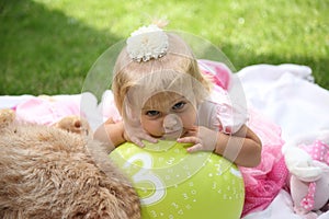 Sweet smiling little girl with long blond hair, sitting on grass in summer park, closeup outdoor portrait.